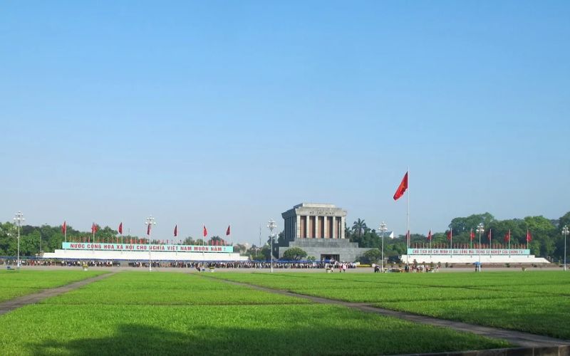 Ho Chi Minh Mausoleum beyond broad green lawns, with Vietnamese flags and visitors under a clear blue sky.