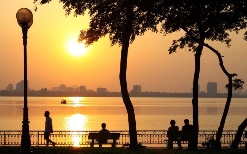 Golden sunset over the West lake in Hanoi, with silhouetted trees, people walking and sitting on waterfront benches, and distant buildings.