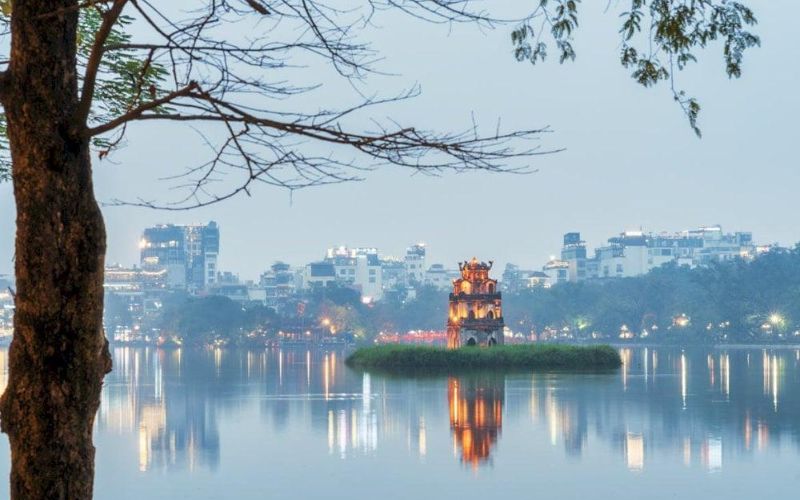 Illuminated Turtle Tower on a small island, reflected in the calm lake at dusk, with tree branches framing the city skyline.