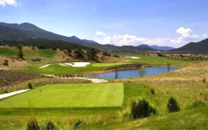 Jade Dragon Snow Mountain Golf Club with a tee box in the foreground, water hazard and fairways in the middle ground, surrounded by rolling hills and mountains under a clear blue sky