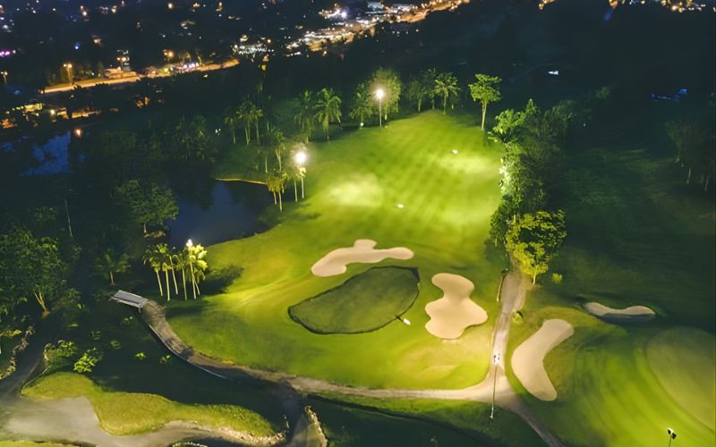 Aerial night view of a brightly illuminated golf hole with sand bunkers and lush green fairways at Tiara Melaka Golf & Country Club, surrounded by dark landscape and distant city lights.