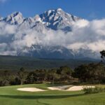 A scenic golf course with a sand bunker in the foreground, set against the dramatic backdrop of Jade Dragon Snow Mountain covered in clouds and snow