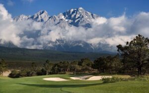 A scenic golf course with a sand bunker in the foreground, set against the dramatic backdrop of Jade Dragon Snow Mountain covered in clouds and snow