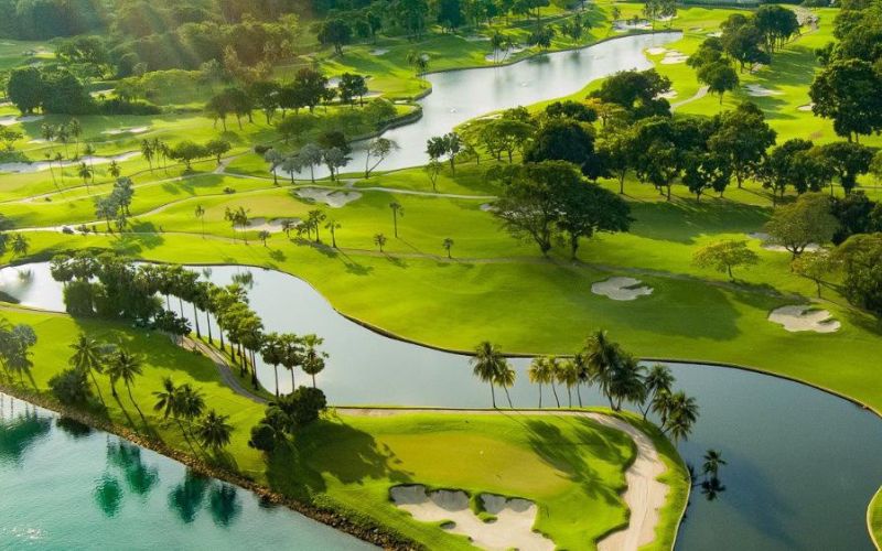 The Sentosa Golf Club in Singapore presents an aerial view, showcasing lush green fairways, water hazards, sand bunkers, and palm trees near the coastline.