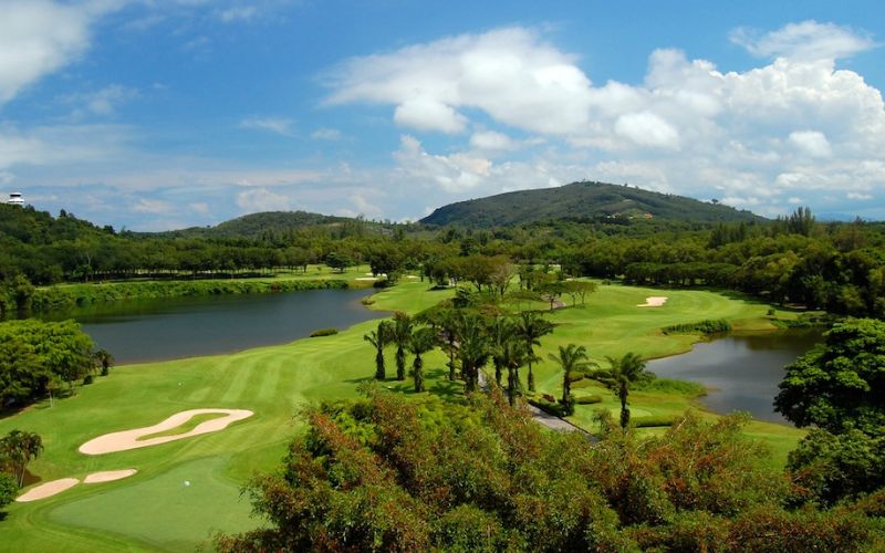 Blue Canyon Country Club in Phuket featuring green fairways, sand bunkers, lakes, and forested hills under a bright sky.