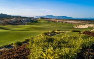 Scenic view of KN Golf Links Cam Ranh featuring coastal fairways, sandy dunes, and a backdrop of mountains under clear blue skies.