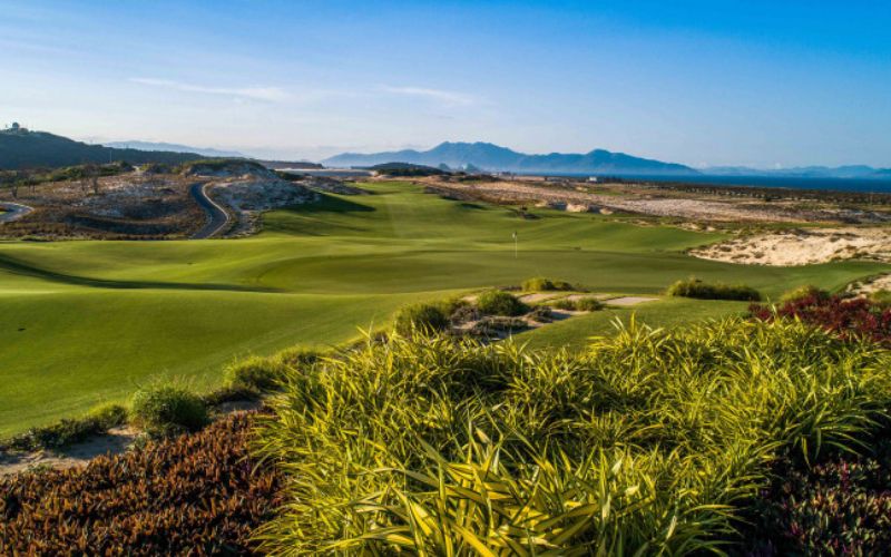 Scenic view of KN Golf Links Cam Ranh featuring coastal fairways, sandy dunes, and a backdrop of mountains under clear blue skies.