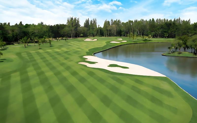 A wide view of a golf course at Muang Kaew Golf Club, showing neatly striped fairways, a sand bunker, and a water hazard, surrounded by trees under a clear sky