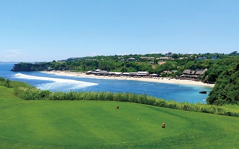 Panoramic view of a golf course with lush green grass, leading to a coastline with clear blue water and a beach in the distance. The sky is bright and clear