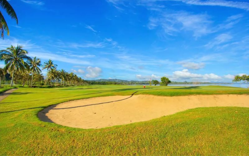 aradise Golf Club course featuring a bright sand bunker, lush green fairway, palm trees, and a vivid blue sky with scattered clouds.