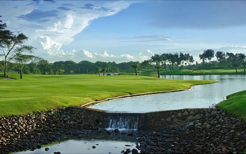 A view of Royale Jakarta Golf Club featuring a well-manicured green, a small water hazard with a waterfall, and trees lining the fairway under a blue sky.