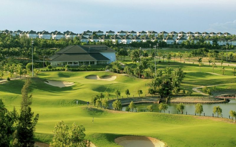 A panoramic view of Sea Links Golf Country Club in Phan Thiet, Vietnam, showing lush green fairways, water features, and a modern clubhouse surrounded by palm trees under a clear sky.