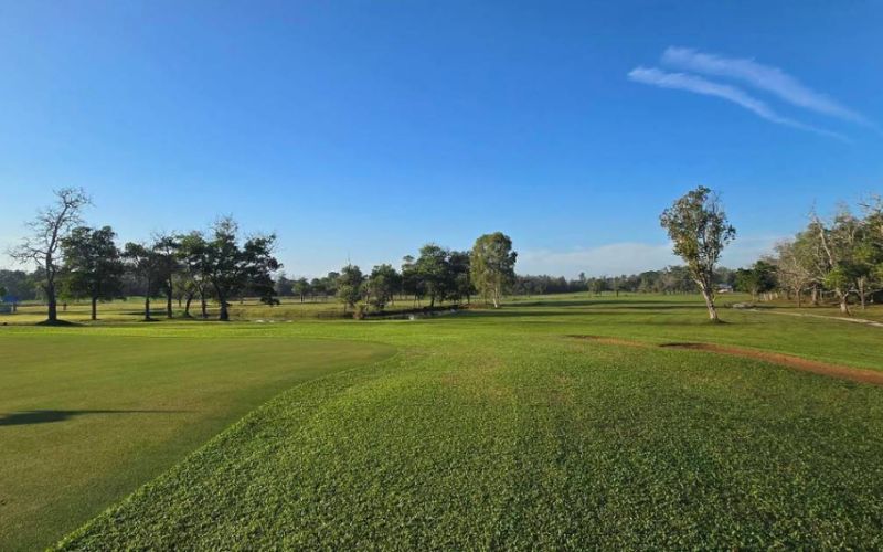 A panoramic view of Swargaloka Golf Club with expansive green fairways, scattered trees, and a clear blue sky.