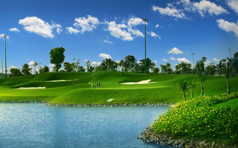 Scenic view of Tan Son Nhat Golf Course with a calm water hazard, manicured green fairways, trees, and a bright blue sky with scattered clouds.