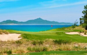 Coastal view of BRG Da Nang Golf Resort featuring green fairways, sandy dunes, and turquoise sea with mountains in the background.