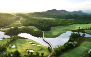 Aerial view of Ba Na Hills Golf Club surrounded by lush mountains, winding fairways, and reflective lakes under warm sunlight.