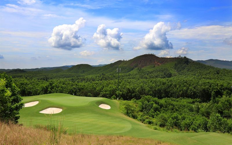 Scenic view of Ba Na Hills Golf Club with green fairways, sand bunkers, and a forested mountain under a bright blue sky with white clouds.