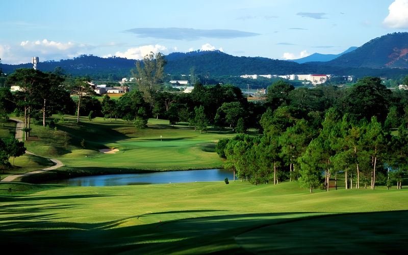 Scenic view of Dalat Palace Golf Club surrounded by pine trees, rolling hills, and mountain landscapes under a clear blue sky.