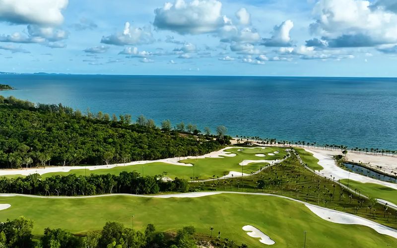 Eschuri Vung Bau Golf Course with green fairways stretching toward the coastline, bordered by palm trees and overlooking the blue ocean under a partly cloudy sky