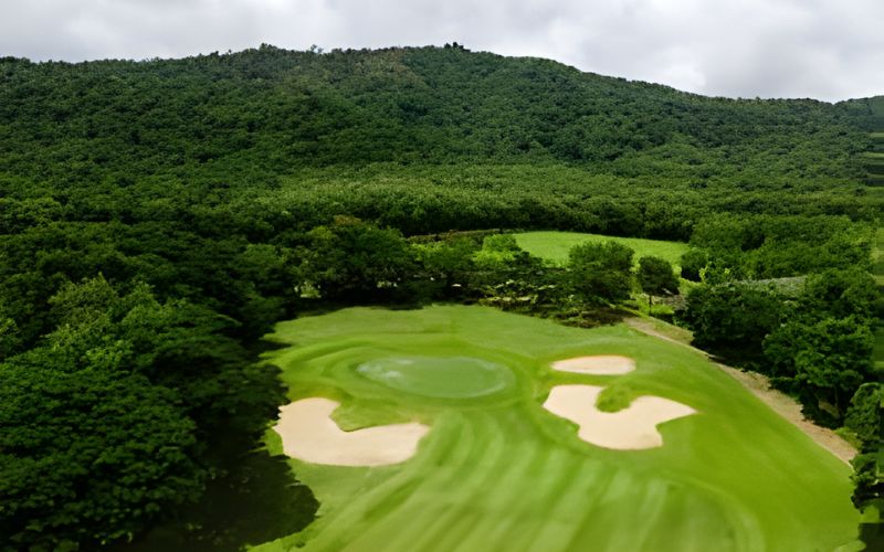 A lush green golf course with sand bunkers surrounded by forested hills under a cloudy sky at Kirimaya Golf Resort.