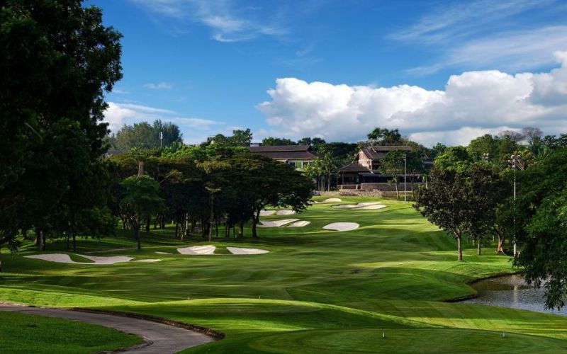 View of Kota Permai Golf & Country Club featuring green fairways, sand bunkers, and a clubhouse surrounded by tall trees under a partly cloudy sky.