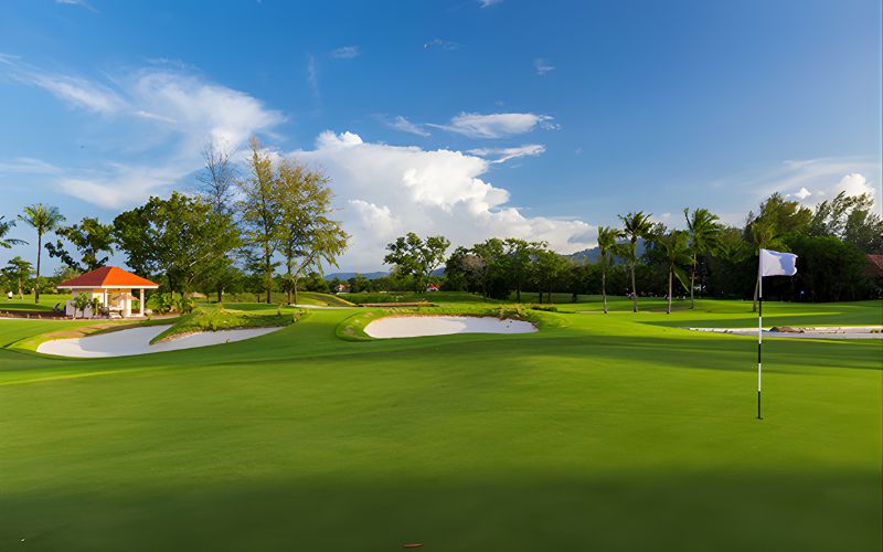 Laguna Golf Phuket with a manicured green, sand bunkers, and a flagstick, surrounded by palm trees and tropical landscaping under a bright blue sky with scattered clouds.