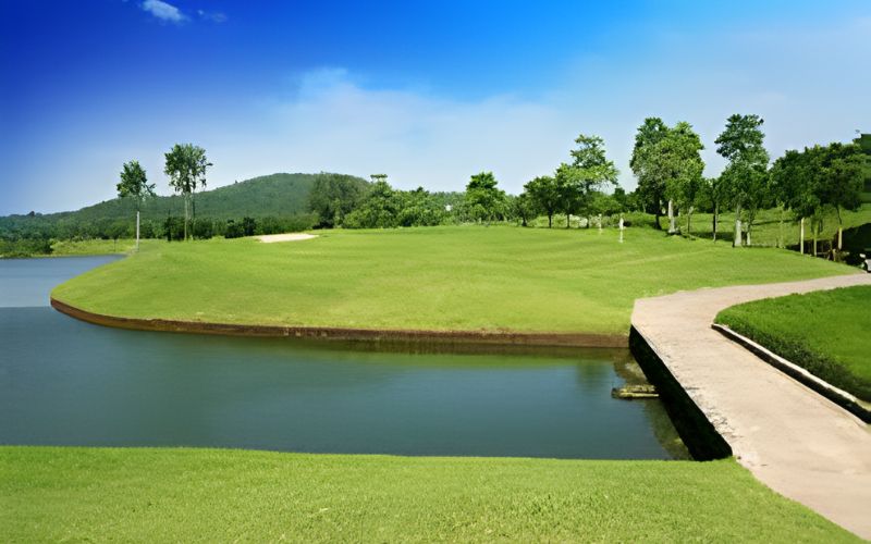 Scenic view of Pattana Golf Club and Resort in Thailand, showcasing a green fairway beside a calm lake with a clear blue sky and lush trees in the background.