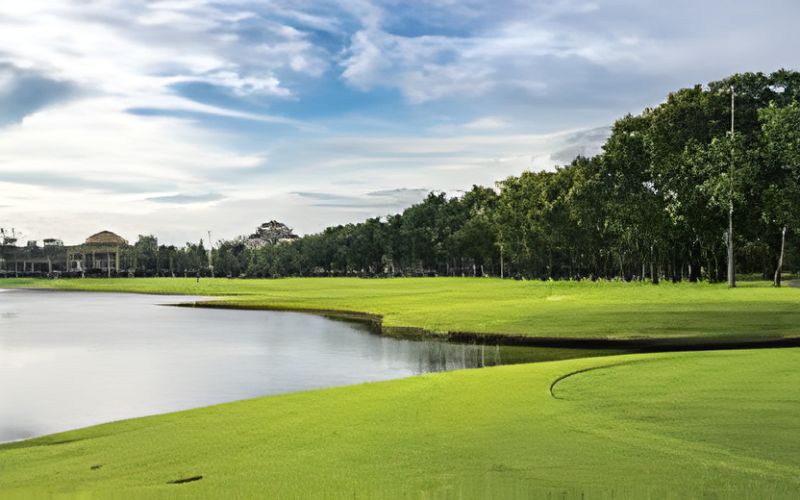 A scenic view of Thana City Country Club golf course in Thailand, featuring lush green fairways beside a calm water hazard under a bright blue sky.