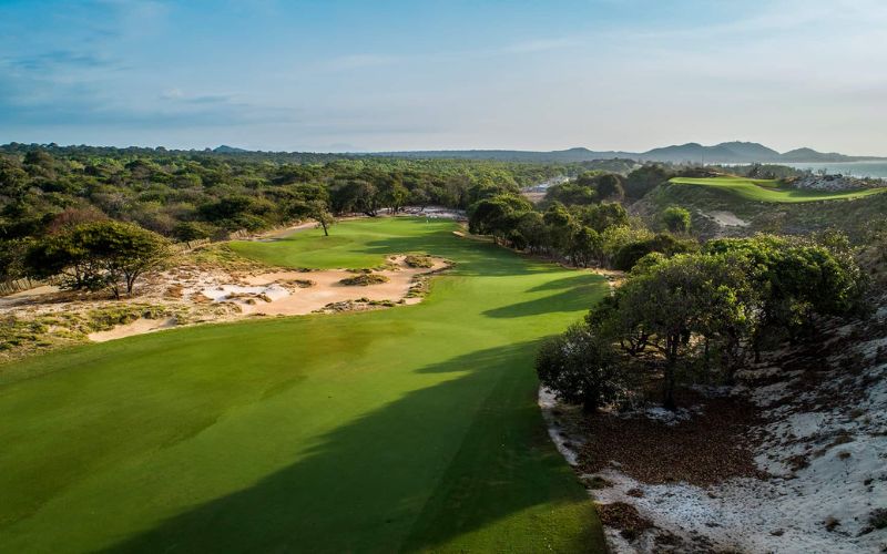 Scenic view of The Bluffs Ho Tram Strip golf course featuring lush green fairways, sand dunes, and surrounding coastal landscape under a clear blue sky.
