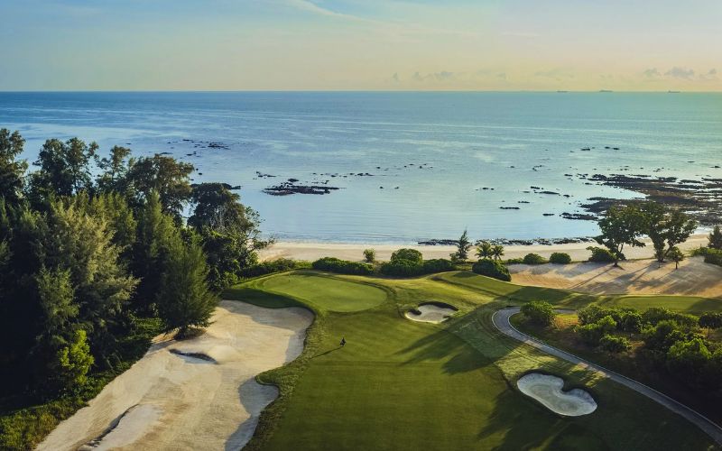 Aerial view of The Els Club Desaru Coast golf course with lush green fairways, sand bunkers, and the ocean in the background at sunrise.