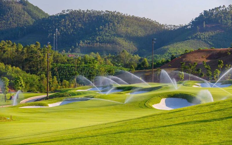 Sprinklers watering the lush green fairway and sand bunkers at Van Lang Empire Golf Club, surrounded by hills and trees under a clear sky.
