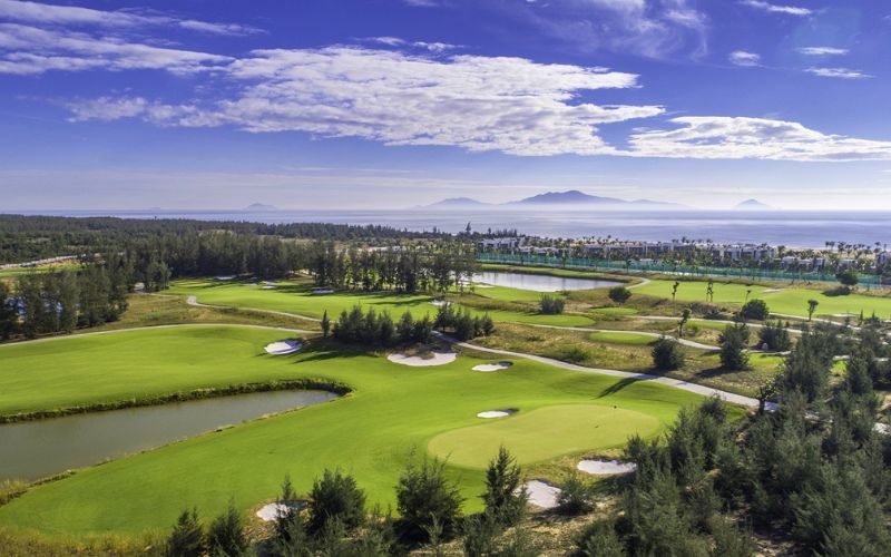 Scenic aerial view of Vinpearl Golf Nam Hoi An featuring lush green fairways, sand bunkers, and a coastal backdrop under a bright blue sky.