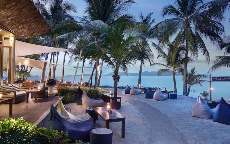 Evening view of Santiburi Koh Samui resort with palm trees, outdoor seating, and the ocean in the background under a twilight sky.
