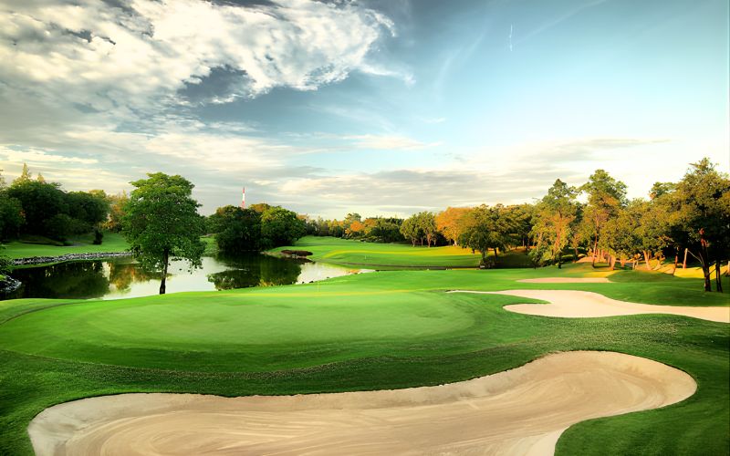 Beautiful view of Alpine Golf Sports Club in Bangkok, Thailand, showcasing a lush green fairway, large sand bunker, and surrounding trees under a soft, glowing sunset sky.