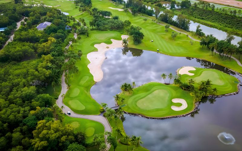 Aerial view of Alpine Golf and Sports Club featuring lush green fairways, a lake surrounding the island-style green, and dense tropical trees under soft daylight.