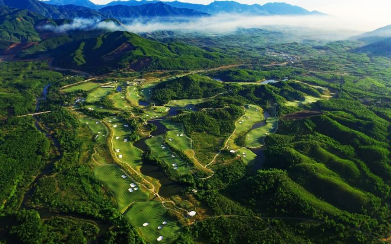 Aerial view of Ba Na Hills Golf Club, showcasing green fairways winding through lush, mountainous terrain under a bright sky.