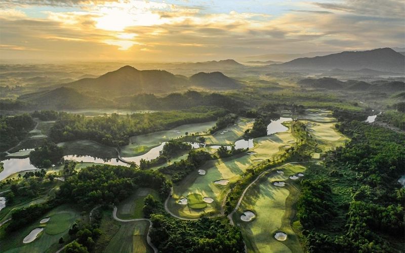 Aerial view of Ba Na Hills Golf Club showing lush green fairways, surrounding trees, and distant mountains under a bright blue sky.