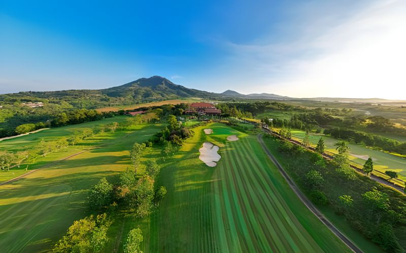 Aerial view of Bandung Giri Golf Resort showing lush green fairways, surrounding trees, and distant mountains under a bright blue sky.