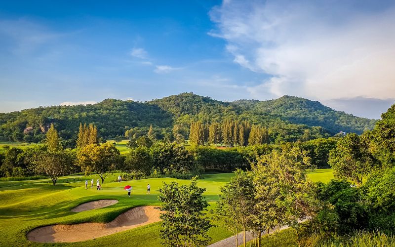 Scenic view of Banyan Golf Club in Hua Hin, featuring lush fairways, sand bunkers, and tree-lined greens set against rolling hills and a bright blue sky.