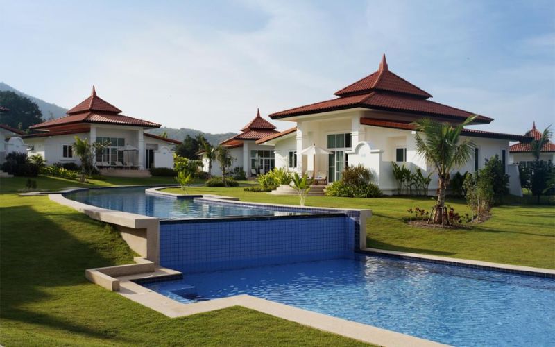 Banyan The Resort Hua Hin featuring tropical-style villas with red-tiled roofs, a clear blue swimming pool, and lush green lawns under a bright sunny sky.