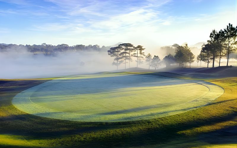 Early morning view of a golf green at Dalat Palace Golf Club with soft mist rising over pine trees and warm sunlight casting long shadows across the fairway.