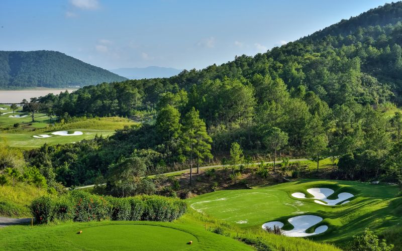 Aerial view of Dalat Palace Golf Club surrounded by rolling hills and pine forests, featuring manicured fairways and bunkers under a bright blue sky.