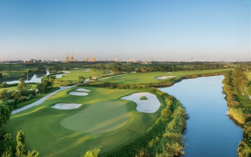 Aerial view of FLC Halong Bay Golf Club showcasing green fairways, sand bunkers, and a winding water feature surrounded by lush landscape under clear blue skies.