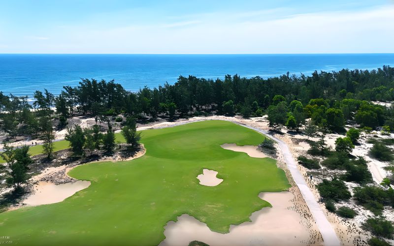 Aerial view of Golden Sands Golf Resort in Vietnam, showing a lush green fairway, sand bunkers, surrounding palm trees, and the blue sea in the background.