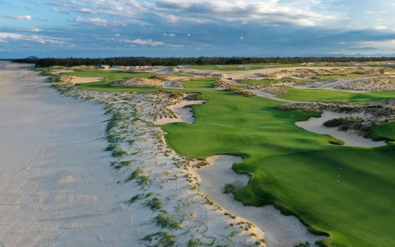 Coastal fairway at Hoiana Shores Golf Club with rolling dunes, sandy edges, and ocean waves running alongside the green under a cloudy sky.