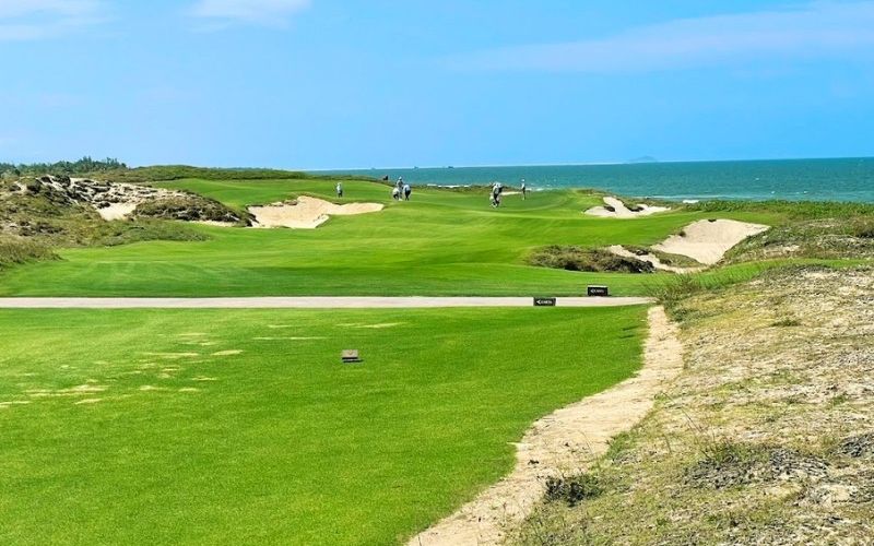 A links-style golf course, likely Hoiana Shores, showing a lush green fairway bordered by large, natural-looking sand dunes and bunkers, with a glimpse of the ocean in the distance.
