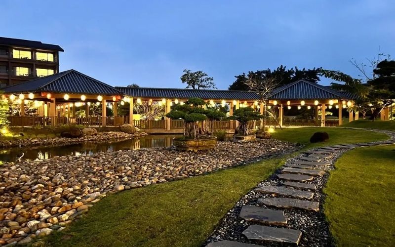 A nighttime view of Kobi Onsen resort Hue with warm lights glowing from wooden buildings, a stone walkway leading through a landscaped garden, and a soft blue sky in the background.