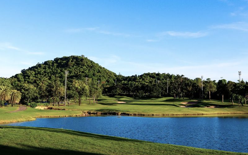 Scenic view of Laem Chabang International Country Club, featuring a lush green golf course with a calm water hazard and forested hills under a clear blue sky.