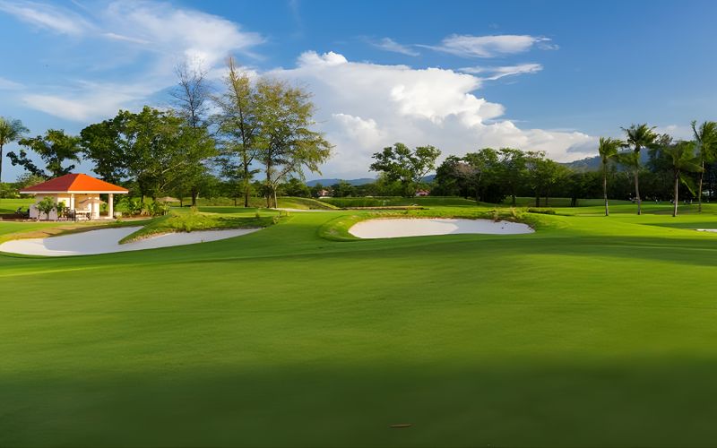 Beautiful view of Laguna Golf Phuket with manicured greens, sand bunkers, lush trees, and a clubhouse under a bright blue sky.