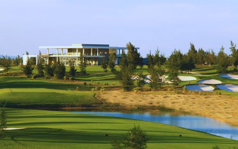 A wide view of Montgomerie Links Vietnam golf course with lush green fairways, a modern clubhouse, sand bunkers, and trees under a clear sky.